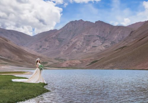 A bride in a wedding dress stands by a tranquil lake in a mountainous valley under a bright sky.