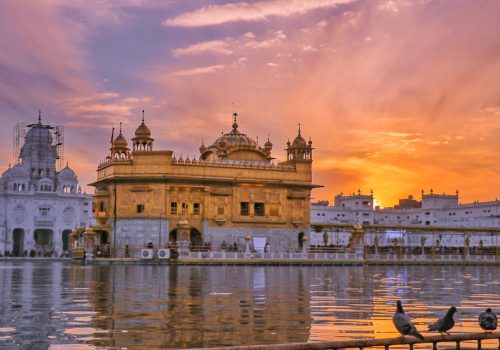 Exterior of Sikh gurdwara golden temple with dome located near water against cloudy sky in evening time in city in India